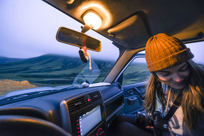 Woman with camera and hat sitting in car looking down at sunset
