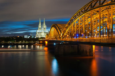 Illuminated bridge over river against buildings at night