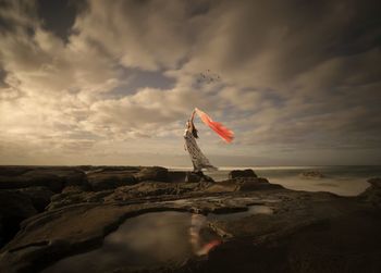 Woman holding scarf standing on rocky shore against cloudy sky during windy weather