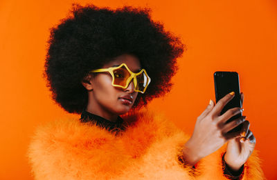 Young woman taking selfie on phone while wearing sunglasses against orange background