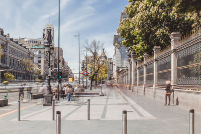 People walking on sidewalk by buildings in city against sky