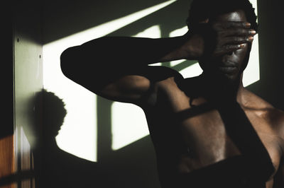 Shirtless man covering eyes while standing in sunlight against wall