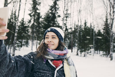 Smiling woman taking selfie through mobile phone while standing against trees on snowy field