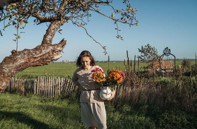 Full length of woman standing on field against sky holding orange flowers