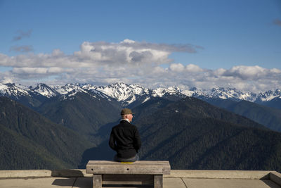 Rear view of man looking at snowcapped mountains while sitting on bench at olympic national park
