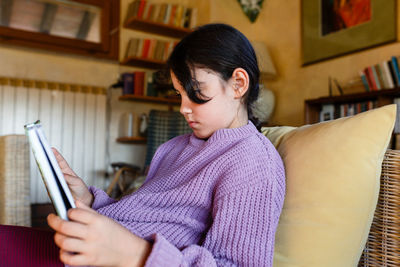 Side view of girl in lilac pullover reading a book in yellow armchair