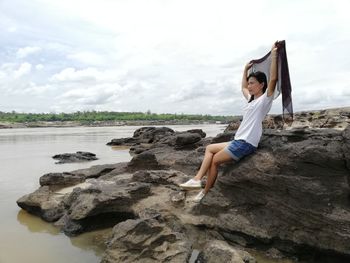 Full length of woman sitting on rock at beach against sky