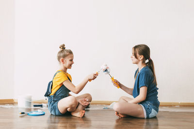 A girl with her sister in bright blue and yellow clothes helps to paint the walls in her room white.