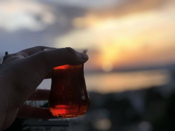 Close-up of hand holding glass bottle against sky during sunset