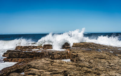 Waves splashing on rocks against clear sky