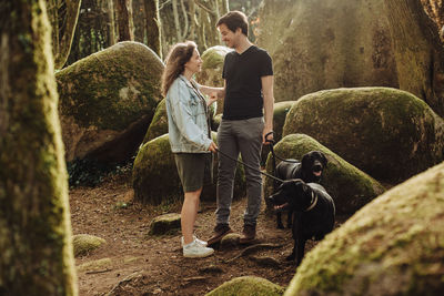 Man and woman smiling to each other while standing with dogs in forest