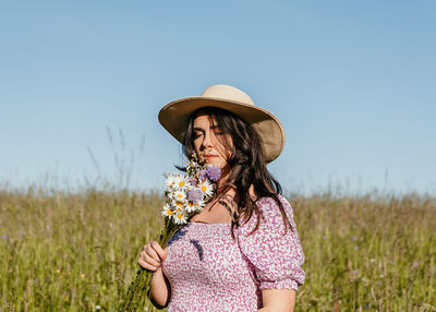 Beautiful woman standing in field, holding and smelling wildflowers