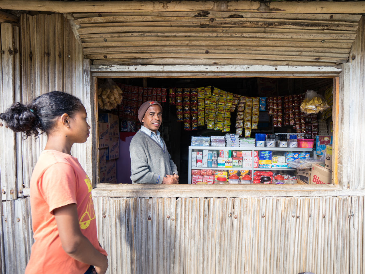 Maubisse, east timor - august 11, 2018: young ethnic female customer approaching to small local grocery store while smiling male vendor standing at counter and looking at camera