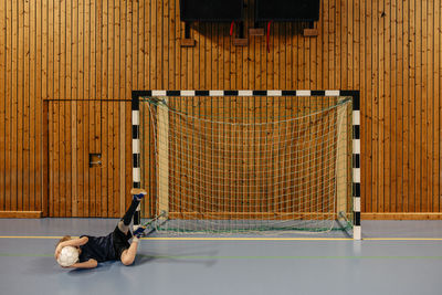 Male goalie with disabled leg lying on floor holding football while playing at sports court