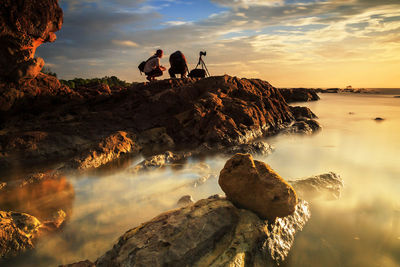 People on rock by sea against sky during sunset