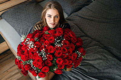 A beautiful girl is sitting on a bed in the bedroom with a huge bouquet of scarlet roses. 