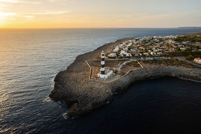 From above amazing remote view of lighthouse located on rocky cliff in front of calm sea near town with white houses in menorca, spain