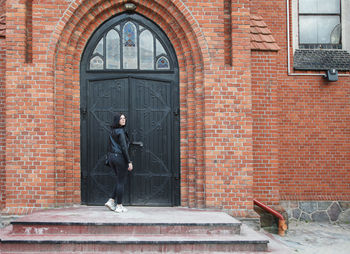 Young beautiful brunette girl in black jacket and jeans stands near door of gothic church on summer