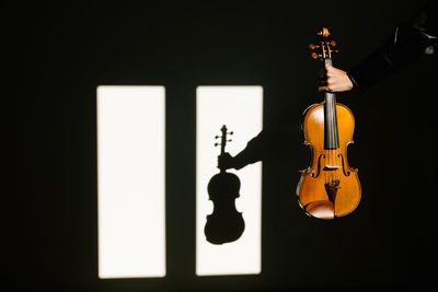 Crop unrecognizable musician in silk black shirt holding modern acoustic violin in outstretched hand in dark room against window on sunny day person