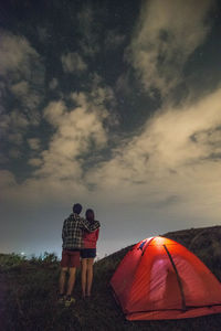 Rear view of man and woman standing by tent on field against cloudy sky at dusk