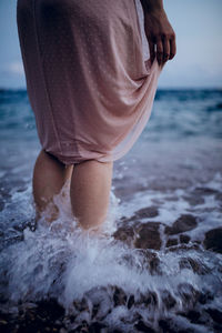 Back view of crop unrecognizable female in pink dress standing in seawater in summer evening
