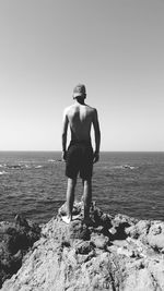 Rear view of shirtless man standing on rock at beach against clear sky