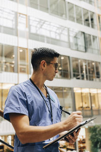 Mature male healthcare worker looking away while writing in clipboard at hospital