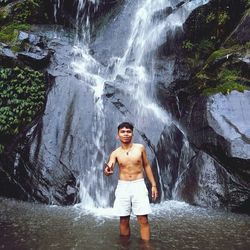 Portrait of shirtless young man gesturing while standing against waterfall