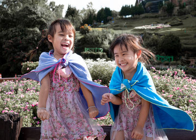 Cheerful smiling girls wearing cape standing against flowering plants