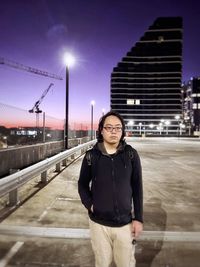 Portrait of young asian man standing on rooftop parking lot against lights and skyscrapers.