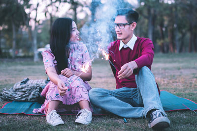 Full length of smiling young woman holding sparklers while sitting on field