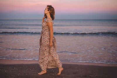 Young woman walking at beach against sky during sunset