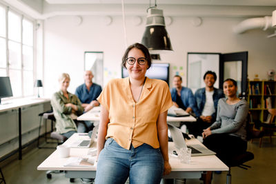 Portrait of smiling female professional leaning on table against executives in modern office