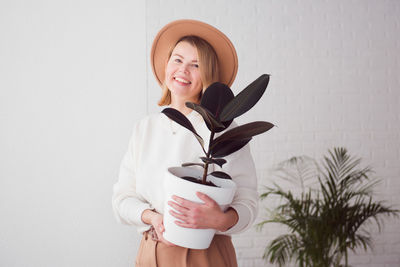 Trendy woman wearing brown hat holding homeplant ficus pot, smiling and looking at camera