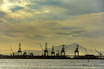Silhouette cranes at harbor against sky during sunset