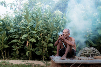 Shirtless senior man smoking while sitting against plants