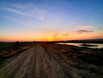 Dirt road amidst field against sky during sunset