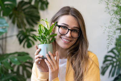 Portrait of woman in glasses holding pot with home plant near face in flower shop or greenhouse