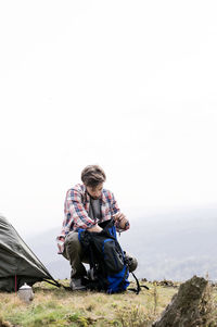 Man searching in backpack at campsite on hill against clear sky