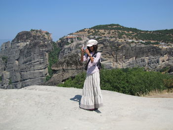 Woman photographing while standing against rock formations against clear blue sky