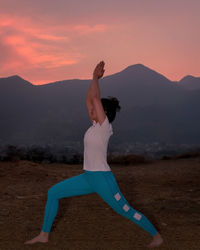 Woman with arms raised standing on land against sky during sunset