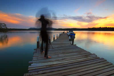 People standing on pier over lake against sky during sunset