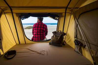 Rear view of man sitting in tent by sea against sky