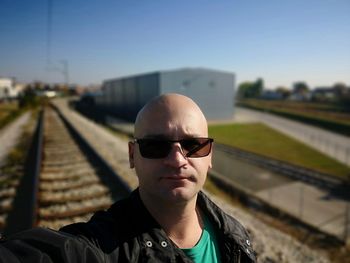 Portrait of bald young man wearing sunglasses at railroad tracks against sky