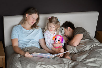 High angle view of mother reading book for daughter while father sleeping on bed at home