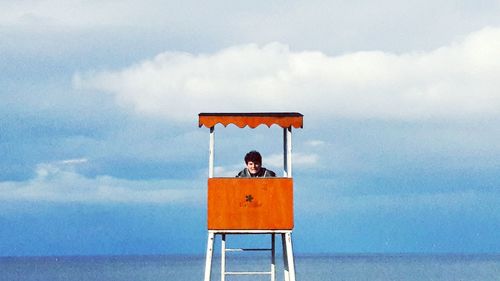 Man sitting at lifeguard hut by sea against sky