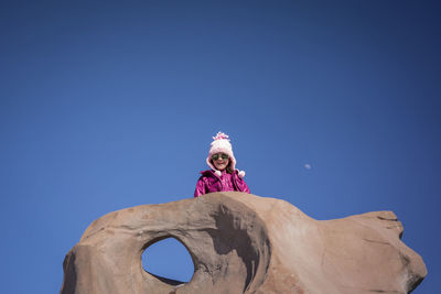 Low angle portrait of happy girl wearing sunglasses while standing on rock formation against clear blue sky during sunny day