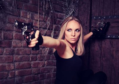Young woman holding gun while crouching against brick wall