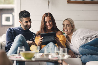 Cheerful young friends sharing digital tablet while sitting on sofa at home