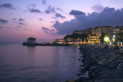 Illuminated buildings by sea against sky at sunset